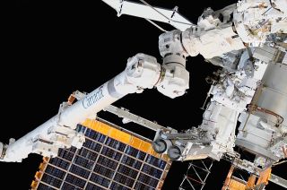 a large robotic arm in front of the space station's solar arrays.  two astronauts are visible nearby in spacesuits