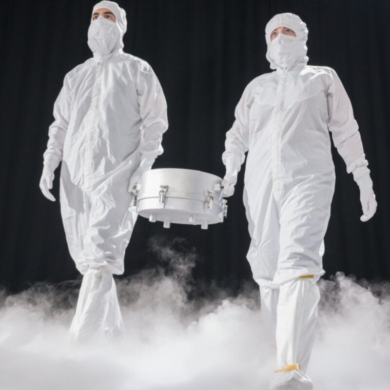 Two men in white suits holding a silver canister between them, in a dark area with white mist billowing from the floor.